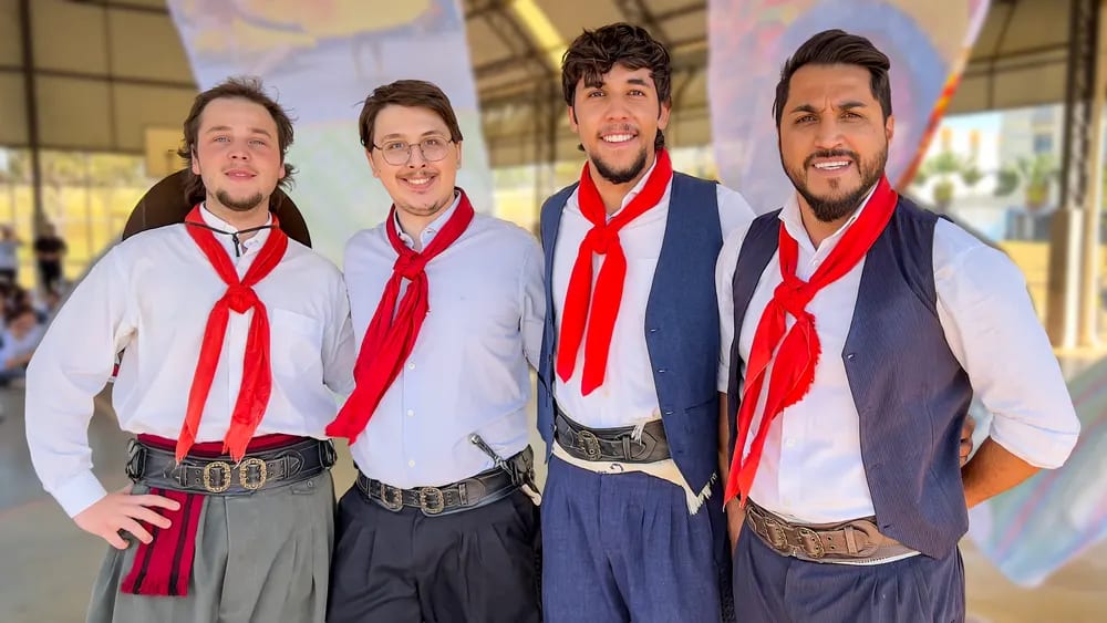 Quatro dançarinos adultos em traje gaúcho — camisa branca, colete e lenço vermelho — posam no pátio da escola